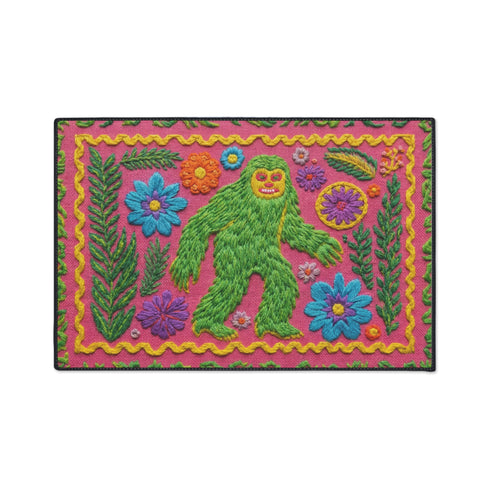 A rectangular flat-printed doormat featuring a faux-tufted, trompe-l'œil texture design. The background is a vibrant hot pink, bordered by a wavy yellow line. In the center is a bright green, smiling Bigfoot or Sasquatch figure with long shaggy hair. Surrounding the cryptid are highly detailed, colorful faux-embroidered floral motifs including blue, orange, and purple daisies, alongside green fern fronds and leaves. The mat is finished with a solid black fabric trim around the outer edge.