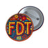 Round pinback metal button featuring an embroidered-style design on a vibrant, multicolored background. The bold, textured letters "FDT" are prominently displayed in red with intricate golden-yellow stitching details, outlined in dark blue. Surrounding the text is an ornate floral pattern with embroidered-style flowers in shades of red, orange, blue, and yellow, densely arranged for a rich, decorative effect. The outer edge is bordered with a zigzag stitching effect in red and blue. The button's back is sil