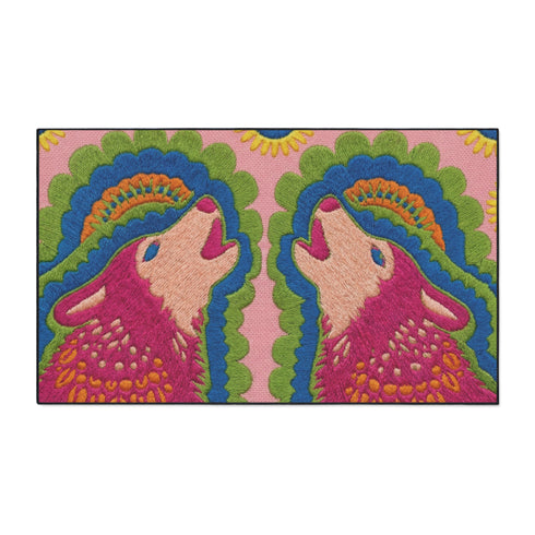 A rectangular outdoor doormat featuring a symmetrical graphic illustration of two howling wolves facing upward in profile. The design utilizes a vibrant psychedelic folk art style with a color palette of magenta pink wolves, peach muzzles, and scalloped layers of lime green and cyan blue foliage. A marigold yellow sun sits at the top center. The background is a dusty rose pink. The texture is a smooth, durable flat 2D print on polyester with a black non-slip rubber border.