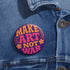A round, glossy purple pinback button with the words "MAKE ART NOT WAR" in a groovy, retro 70s-style font. The word "MAKE" is in orange, "ART" is in a vibrant pink, and "NOT WAR" is in orange. The text is surrounded by small, stylized flower and star details. This psychedelic protest pin is a piece of activist merch with a strong metal back and safety pin clasp, perfect as an anti-war or social justice accessory for a jean jacket or backpack. The pin has a handmade, indie aesthetic.