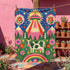 A rectangular velveteen minky blanket with a bright, maximalist design. The top half features a vibrant blue sky with a pink, green, and yellow UFO beaming rays down over hot pink snow-capped mountains. The bottom half shows a grassy green field with a white cow covered in dark spots, surrounded by large, colorful red, pink, and blue retro flowers, pine trees, and a rainbow arch at the very bottom. The reverse side is a fluffy off-white sherpa.