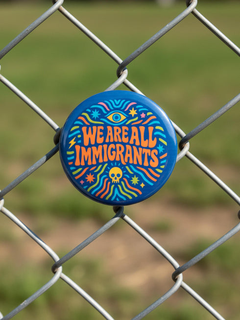 A round 1.75 inch blue pinback button featuring retro psychedelic artwork. The text reads We Are All Immigrants in a groovy orange and red 1970s font style centered on the badge. A mystic third eye sits above the text while a small skull sits below it. Wavy rainbow lines in cyan, green, and orange radiate outward from the center along with small starbursts and lightning bolts. The button has a glossy finish reflecting light.