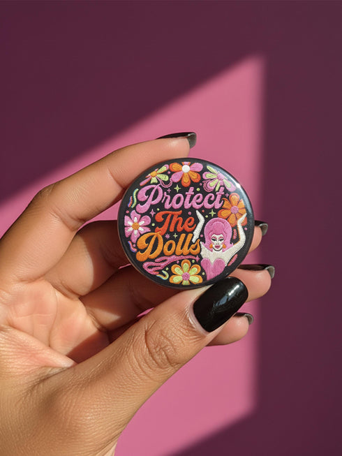 A circular black pinback button featuring the slogan Protect The Dolls in retro script. The design utilizes a high-fidelity trompe-l'œil print style that mimics the texture of raised thread embroidery with satin-stitch detailing. The artwork features vibrant hot pink, rust orange, and golden yellow motifs including drag queens with bouffant hair, 70s style daisies, lightning bolts, and a crescent moon with a face. The surface is smooth and glossy, creating a faux-textured illusion.