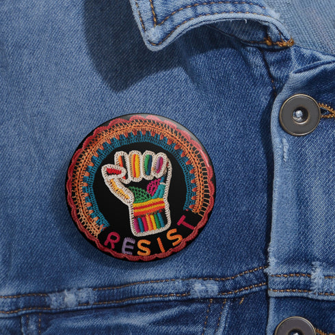 Round pinback metal button with an embroidered-style design on a black background. The center features a raised fist symbol stitched in a multicolored pattern, incorporating rainbow hues and folk-inspired textures. The fist is encircled by a vibrant, lace-like border in shades of blue, orange, and red, resembling intricate embroidery. Below the fist, the word "RESIST" is spelled out in bold, stitched-textured letters in red and orange. The button has a silver metal back with a secure pin closure.