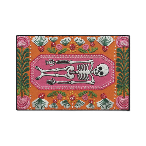A rectangular terracotta orange doormat featuring a central illustration of a white skeleton lying inside a pink coffin shape. The design is surrounded by a symmetrical pattern of green leaves, pink flowers, and red and white spotted mushrooms. The imagery uses a trompe-l'œil effect to mimic the texture of raised needlepoint embroidery, but the surface is flat printed polyester. The mat is framed with a solid black edge.