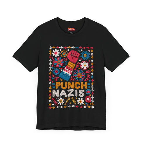 Black t-shirt featuring an embroidered-style design with the phrase "PUNCH NAZIS" in bold, colorful lettering. Above the text, a raised red fist is illustrated, symbolizing resistance, and is surrounded by a vibrant folk-art-inspired floral border with bright flowers, leaves, and geometric patterns in red, yellow, blue, and white.