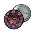 Embroidered-style pinback button featuring the phrase "POWER TO THE PEOPLE" in bold, multicolored satin-stitch letters—green, purple, and orange—framed by a vivid circular floral wreath. Pink, red, and purple flowers with leafy greens radiate around the text. Design has a handcrafted, folk-art feel with a transparent background.