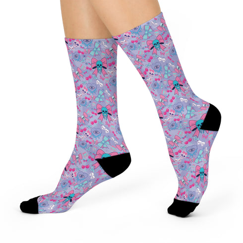 Knee-high socks worn on legs, featuring a pastel blue background with a quirky pattern. The pattern includes pink and teal skulls, some adorned with bows, alongside images of cherries, hearts, and additional bow designs. The socks have black toes and heels, contrasting with the colorful pattern. The overall effect is a playful mix of cute and edgy elements.