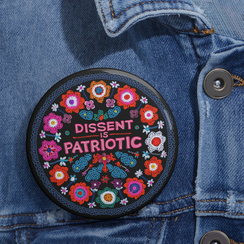 Round black pinback button with vibrant floral embroidery design surrounding bold pink and red text reading "Dissent is Patriotic."