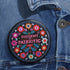 Round black pinback button with vibrant floral embroidery design surrounding bold pink and red text reading "Dissent is Patriotic."
