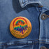 Round orange pinback button featuring bold rainbow psychedelic artwork of a melting third-eye face beneath a rainbow arch. Surrounded by stars and the phrase “Hot, Queer and Radiating” in retro black bubble letters. Glossy metal badge with UV-resistant mylar face and secure safety pin back.