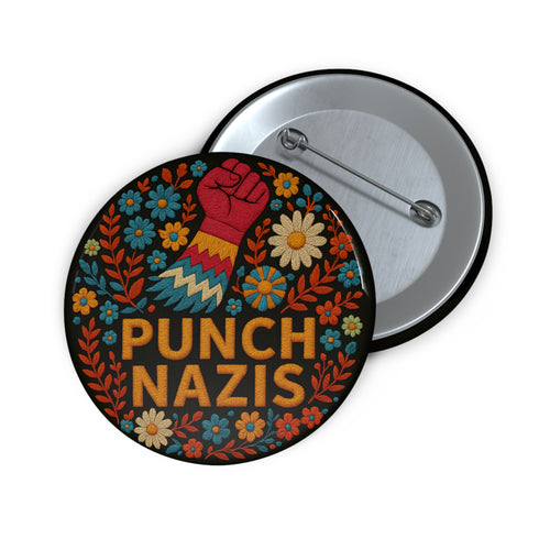 Round black pinback button with bold embroidered-style design featuring a raised red fist, zigzag sleeve, and colorful folk art flowers in orange, teal, yellow, and white. Center text reads “PUNCH NAZIS” in bright golden thread-style lettering. The design is bold, anti-fascist, and maximalist, shown on a glossy pin surface against a pink and purple gradient background. Statement accessory for protest fashion, punk style, and political activism.