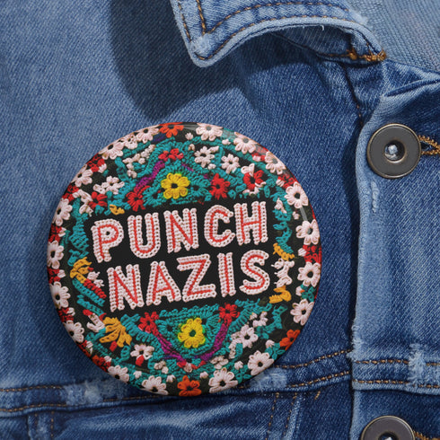 Round pinback metal button with an embroidered-style design on a black background. The center displays the phrase "PUNCH NAZIS" in bold, white stitched-textured lettering with red accents. Surrounding the text is an elaborate folk-inspired floral pattern, densely packed with embroidered-style flowers in shades of red, yellow, blue, pink, and white. The intricate design includes symmetrical lace-like stitching and decorative motifs, creating a rich, tapestry-like effect.