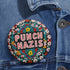 Round pinback metal button with an embroidered-style design on a black background. The center displays the phrase "PUNCH NAZIS" in bold, white stitched-textured lettering with red accents. Surrounding the text is an elaborate folk-inspired floral pattern, densely packed with embroidered-style flowers in shades of red, yellow, blue, pink, and white. The intricate design includes symmetrical lace-like stitching and decorative motifs, creating a rich, tapestry-like effect.