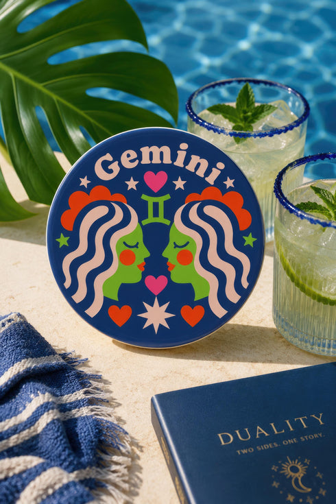 A close-up photograph of a round, glossy ceramic coaster featuring a bold retro pop art design. The background is a deep royal blue. In the center are two symmetrical profile faces of twins looking at each other, featuring bright green skin, closed eyes, and red cheeks. They have wavy white and pale pink hair with red accents. The word Gemini is arched at the top in a retro white font. The design is decorated with a green Gemini symbol, pink and red hearts, and white starbursts.