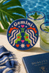 A close-up photograph of a round, glossy ceramic coaster featuring a bold retro pop art design. The background is a deep royal blue. In the center are two symmetrical profile faces of twins looking at each other, featuring bright green skin, closed eyes, and red cheeks. They have wavy white and pale pink hair with red accents. The word Gemini is arched at the top in a retro white font. The design is decorated with a green Gemini symbol, pink and red hearts, and white starbursts.