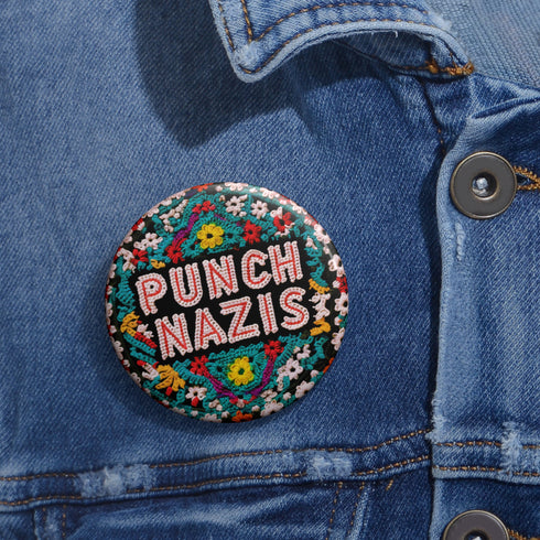 Round pinback metal button with an embroidered-style design on a black background. The center displays the phrase "PUNCH NAZIS" in bold, white stitched-textured lettering with red accents. Surrounding the text is an elaborate folk-inspired floral pattern, densely packed with embroidered-style flowers in shades of red, yellow, blue, pink, and white. The intricate design includes symmetrical lace-like stitching and decorative motifs, creating a rich, tapestry-like effect.