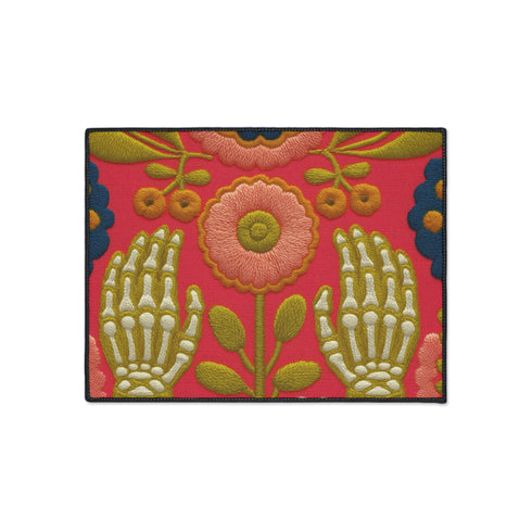 Pink outdoor doormat featuring a symmetrical folk art design. Two white skeleton hands with palms facing forward frame a large central pink and peach flower with a yellow center. Olive green stems, gold berries, and teal blue floral accents decorate the vibrant magenta background. The design mimics a textured embroidery look but is a flat 2d print. Framed with a sleek black border.