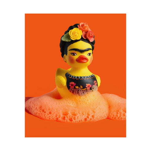 A vibrant fine art print of a rubber duck inspired by Frida Kahlo, complete with bold unibrow, floral crown, and traditional embroidered blouse. Framed in a red sculptural frame, the duck floats in orange-tinted bubbles against a deep orange backdrop and violet wall.