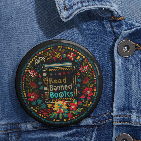 Round black pinback button with vibrant Mexican folk art-inspired embroidery design featuring red, yellow, blue and teal flowers surrounding a gold book spine with "Read Banned Books" text. Literary protest accessory with glossy finish and safety pin backing, available in 2" and 3" sizes.