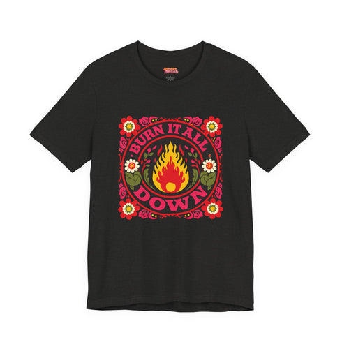A high-resolution product image of a black unisex t-shirt featuring the ‘BURN IT ALL DOWN’ design in a folk-art style. The central motif is a vibrant orange and yellow flame surrounded by a circular medallion and a square floral border with red and pink flowers, white daisies, and green leaves. The print uses a faux-embroidery technique to create a trompe-l’œil 3D texture on a smooth 2D surface. The shirt has a ribbed knit collar, set against a high-contrast background.