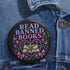 Glossy black pinback button that says “Read Banned Books” in purple and teal letters, featuring a chained book, roses, bows, and floral accents, styled on colorful books and pastel backgrounds.