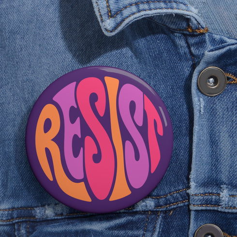 Close-up of a glossy, round purple pinback button. The button features the word "RESIST" in a bold, 70s-style psychedelic font that fills the entire surface. The bubbly, wavy letters alternate in color between a vibrant orange, a light peach-pink, and a deep magenta, creating a powerful and retro statement piece. The design has a distinct groovy and feminist protest aesthetic, making it a perfect accessory for jackets, bags, or activist apparel.