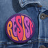 Close-up of a glossy, round purple pinback button. The button features the word "RESIST" in a bold, 70s-style psychedelic font that fills the entire surface. The bubbly, wavy letters alternate in color between a vibrant orange, a light peach-pink, and a deep magenta, creating a powerful and retro statement piece. The design has a distinct groovy and feminist protest aesthetic, making it a perfect accessory for jackets, bags, or activist apparel.