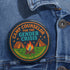Round glossy pinback button measuring 2.25 inches featuring a playful camp-themed design with the phrase “Camp Counselor for the Gender Crisis.” The artwork includes two orange tents, a rainbow campfire, stars, and green grass. Bottom text reads “Cabin Queer, Unit Psychedelic.” Made from durable tinplate with a safety pin back and coated in a UV and scratch-resistant mylar finish. Bold and colorful, perfect for jackets, bags, or pride outfits.