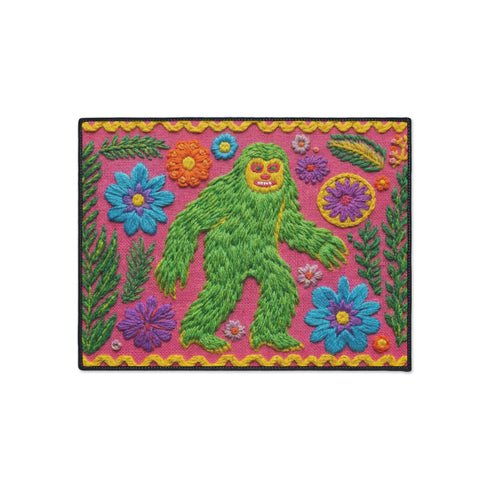 A rectangular flat-printed doormat featuring a faux-tufted, trompe-l'œil texture design. The background is a vibrant hot pink, bordered by a wavy yellow line. In the center is a bright green, smiling Bigfoot or Sasquatch figure with long shaggy hair. Surrounding the cryptid are highly detailed, colorful faux-embroidered floral motifs including blue, orange, and purple daisies, alongside green fern fronds and leaves. The mat is finished with a solid black fabric trim around the outer edge.