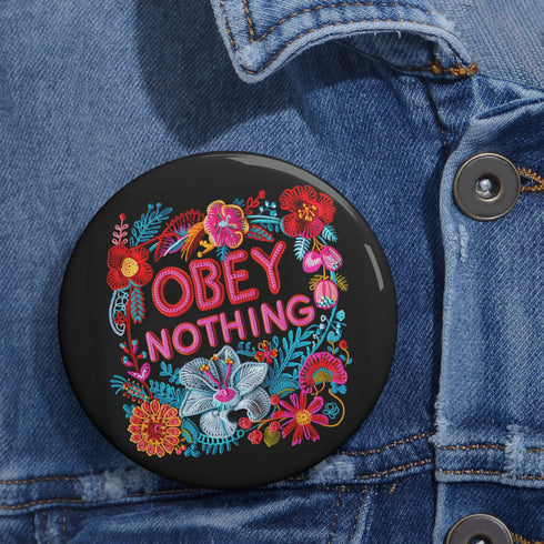 A black round pinback button featuring floral embroidery-style designs and bold red text that reads "Obey Nothing."
