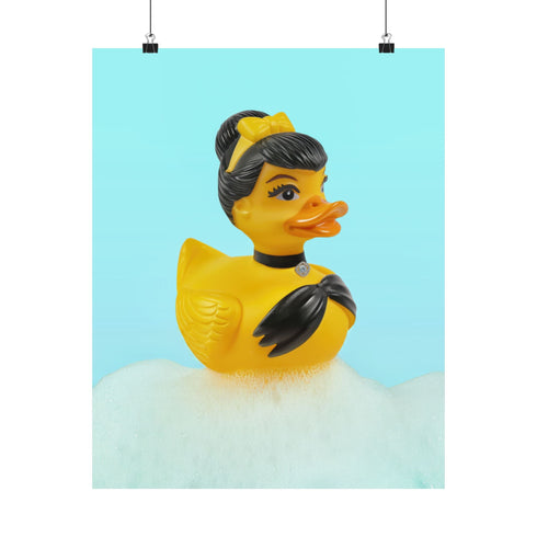 A chic rubber duck styled like Audrey Hepburn’s Holly Golightly, wearing a high bun, yellow bow, black gloves, and a sparkly choker. The duck floats in a pile of foam and is framed in white on a bubbly bathroom shelf with a red tile backdrop and soap dispenser.