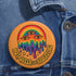 Round orange pinback button featuring bold rainbow psychedelic artwork of a melting third-eye face beneath a rainbow arch. Surrounded by stars and the phrase “Hot, Queer and Radiating” in retro black bubble letters. Glossy metal badge with UV-resistant mylar face and secure safety pin back.