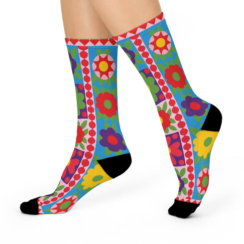 A pair of bright crew socks featuring a vibrant 70s-style maximalist patchwork design. The 2D flat print includes a collage of bold floral motifs in purple, lime green, red, and yellow set against alternating sky blue and light pink square panels. Features a red and white sawtooth border at the cuff and a vertical red scalloped stripe. Reinforced solid black toe and heel. High-quality accessory from black owned shops.