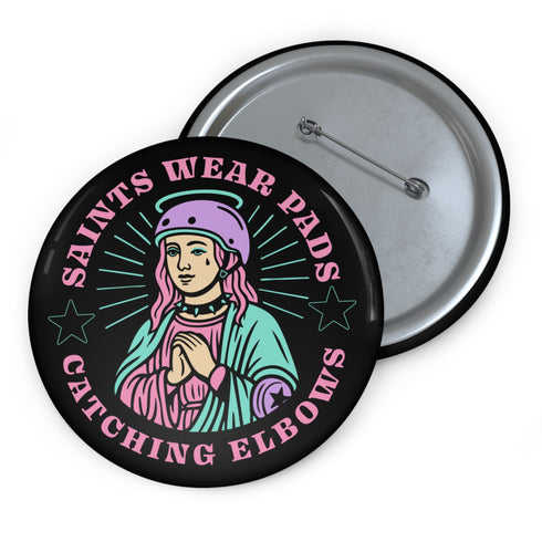 Pinback button badge featuring a saint-inspired roller skater wearing pads and a helmet, with pink hair and teal robes. Surrounded by retro starbursts, the black button reads “Saints Wear Pads Catching Elbows” in pastel pink and teal lettering. Design is bold, colorful, and printed on a glossy metal surface with a classic safety pin back. Available in 2.25" or 3" sizes.