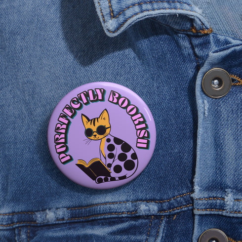 Purple button pin featuring a yellow cat wearing sunglasses and reading a book, with "Purrfectly Bookish" text curved around the edge