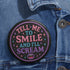 Glossy black pinback button with pastel-colored embroidered-style text that reads “Tell Me to Smile and I’ll Scream,” with lips icon and star accents.