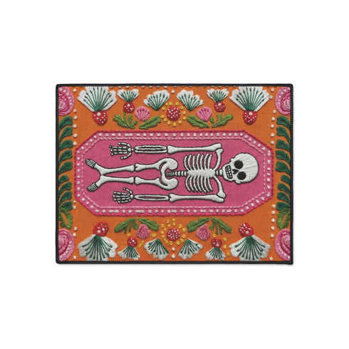 A rectangular terracotta orange doormat featuring a central illustration of a white skeleton lying inside a pink coffin shape. The design is surrounded by a symmetrical pattern of green leaves, pink flowers, and red and white spotted mushrooms. The imagery uses a trompe-l'œil effect to mimic the texture of raised needlepoint embroidery, but the surface is flat printed polyester. The mat is framed with a solid black edge.