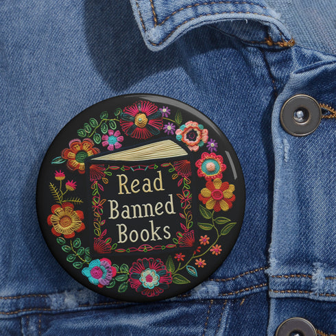 Round black pinback button with "Read Banned Books" in gold lettering on an open book image, surrounded by vibrant embroidery-style flowers in pink, orange, blue and green. Folk art-inspired literary protest pin with glossy finish and safety pin backing, available in 2" and 3" sizes.