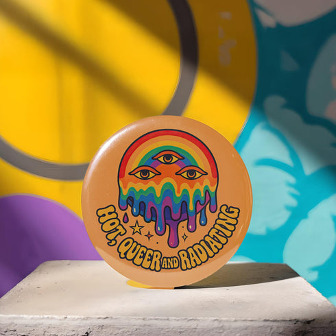 Round orange pinback button featuring bold rainbow psychedelic artwork of a melting third-eye face beneath a rainbow arch. Surrounded by stars and the phrase “Hot, Queer and Radiating” in retro black bubble letters. Glossy metal badge with UV-resistant mylar face and secure safety pin back.