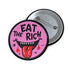 Round pink pinback button featuring a cartoon-style open mouth with sharp teeth, a red tongue, and dripping red details. The bold black text reads “EAT THE RICH” in a hand-drawn style. Glossy black rim with a vibrant, rebellious graphic design.