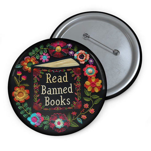 Round black pinback button with "Read Banned Books" in gold lettering on an open book image, surrounded by vibrant embroidery-style flowers in pink, orange, blue and green. Folk art-inspired literary protest pin with glossy finish and safety pin backing, available in 2" and 3" sizes.