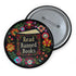 Round black pinback button with "Read Banned Books" in gold lettering on an open book image, surrounded by vibrant embroidery-style flowers in pink, orange, blue and green. Folk art-inspired literary protest pin with glossy finish and safety pin backing, available in 2" and 3" sizes.