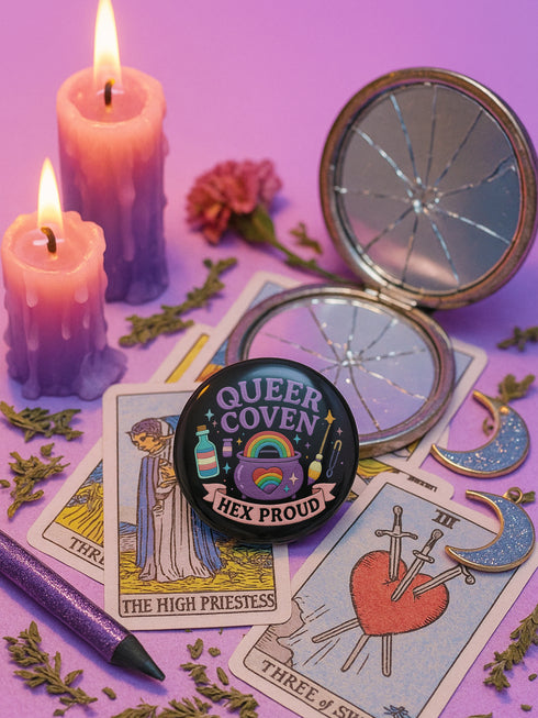 Glossy black pinback button with pastel artwork of a bubbling cauldron, rainbow, potion bottles, and tools. Text reads “Queer Coven, Hex Proud.”