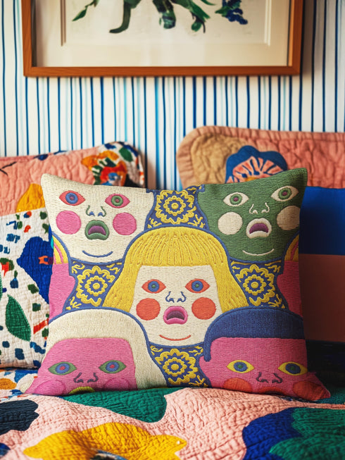A square decorative pillow cover featuring a faux embroidery pattern of five stylized, expressive doll faces. The central face has yellow hair and pink cheeks. It is surrounded by four other faces in shades of pale beige, green, and bright pink, all with wide staring eyes and open mouths. The design uses intricate, colorful line work to create the illusion of thick woven stitching, but the fabric is a smooth print. Medallions fill the gaps.