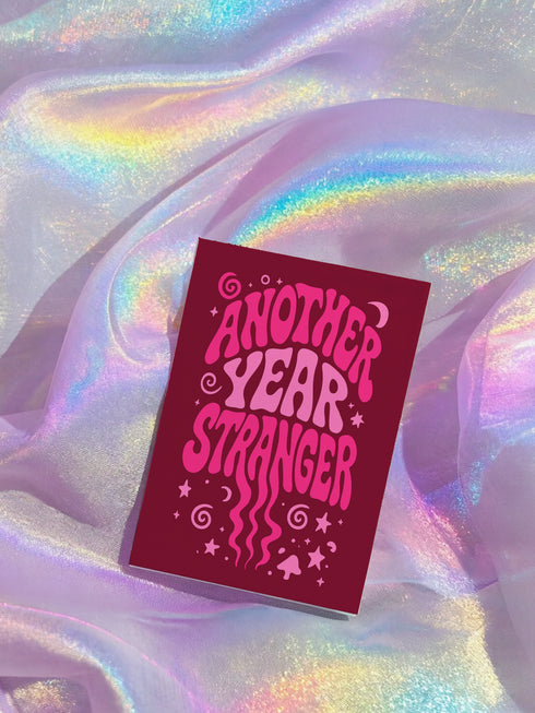 A red book titled "Another Year Stranger" is placed on a shimmering fabric background.
