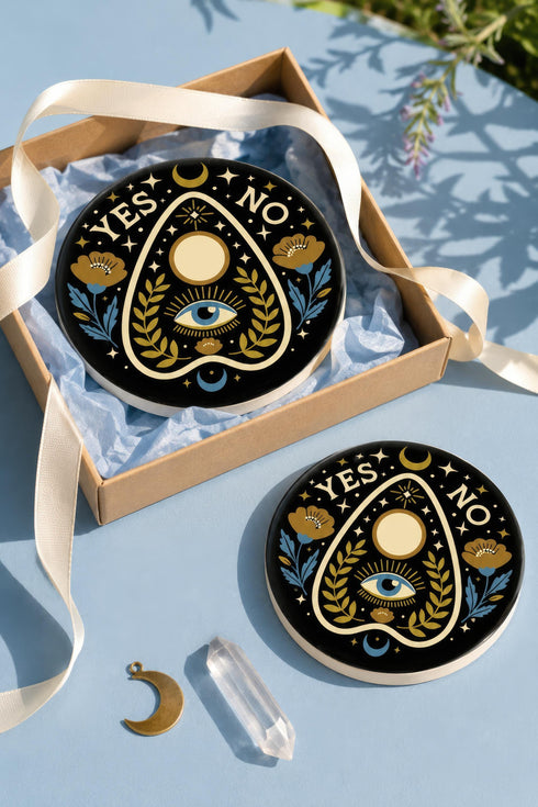 A close-up photograph of a glossy ceramic coaster featuring a Planchette occult design on a deep black background. The symmetrical graphic displays a central blue All-Seeing Eye with gold sunburst lines, topped by a white circle, and flanked by gold flowers with blue stems. At the top, the text "YES" is printed in gold on the left and "NO" on the right. Gold stars and crescent moons are scattered around the perimeter. The flat, round coaster has a highly visible high-gloss glaze.