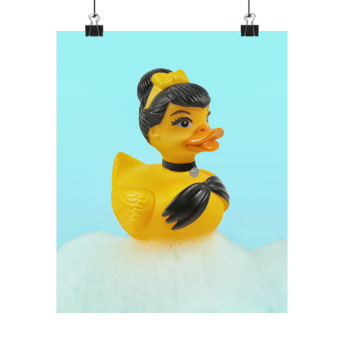 A chic rubber duck styled like Audrey Hepburn’s Holly Golightly, wearing a high bun, yellow bow, black gloves, and a sparkly choker. The duck floats in a pile of foam and is framed in white on a bubbly bathroom shelf with a red tile backdrop and soap dispenser.