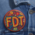 Round pinback metal button featuring an embroidered-style design on a vibrant, multicolored background. The bold, textured letters "FDT" are prominently displayed in red with intricate golden-yellow stitching details, outlined in dark blue. Surrounding the text is an ornate floral pattern with embroidered-style flowers in shades of red, orange, blue, and yellow, densely arranged for a rich, decorative effect. The outer edge is bordered with a zigzag stitching effect in red and blue. The button's back is sil