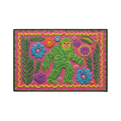 A rectangular flat-printed doormat featuring a faux-tufted, trompe-l'œil texture design. The background is a vibrant hot pink, bordered by a wavy yellow line. In the center is a bright green, smiling Bigfoot or Sasquatch figure with long shaggy hair. Surrounding the cryptid are highly detailed, colorful faux-embroidered floral motifs including blue, orange, and purple daisies, alongside green fern fronds and leaves. The mat is finished with a solid black fabric trim around the outer edge.